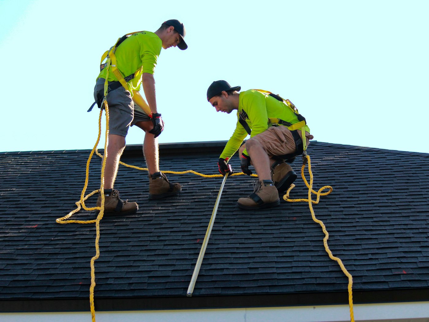 Two roofers in harnesses and hi-vis measuring an asphalt shingle roof